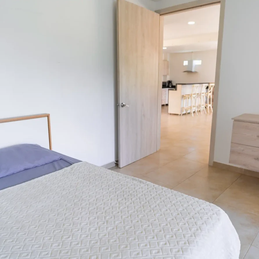 Dormitorio moderno con cama, cómoda de madera y acceso a cocina equipada, ideal para alquiler en Calima El Darién.